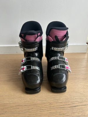 Rossignol svarta pjäxor med rosa detaljer - Säljer ett par svarta pjäxor från Rossignol med rosa och vita detaljer. De har fyra spännen i metall och plast för bra passform och en mjuk, vadderad insida. Perfekta för dig som vill ha snygga och funktionella pjäxor till skidbacken.