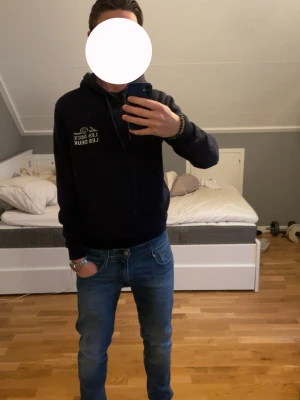 Mörkblå hoodie från Les Deux - Snygg mörkblå hoodie från Les Deux med vit logga tryckt på bröstet. Klassisk modell med huva och dragsnören samt en stor magficka framtill. Tillverkad i mjukt material som känns skönt mot huden. Perfekt för en avslappnad och stilren look. Passar dig mellan 170-180 cm