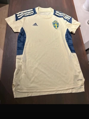 Sverige fotbollströja - Snygg landslagströja från Adidas med blå detaljer och Sveriges landslagsemblem på bröstet. Tröjan har korta ärmar, rund hals och klassiska Adidas-ränder på axlarna. Tillverkad i lätt och ventilerande material med 'Primeblue'-teknologi. Något gulaktig i färgen. Känn dig fri att pruta om priset inte passar