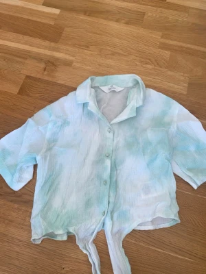 Ljusgrön kortärmad skjorta med knyt - Säljer en luftig och somrig skjorta från H&M i ljusgrönt och vitt. Skjortan har korta ärmar, knappar framtill och en snygg knytning nedtill. Perfekt för varma dagar och enkel att styla till olika outfits. En pytteliten fläck, men som absolut inte syns när man har den på sig!! Se bild 5.