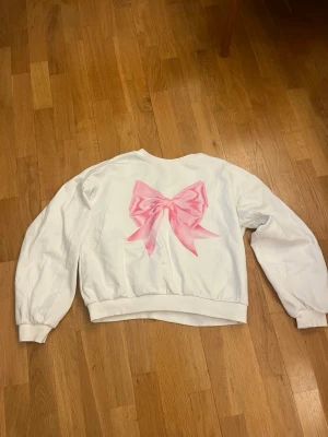 Vit sweatshirt med rosa rosett - Supersöt vit sweatshirt med en stor rosa rosett tryckt på framsidan. Tröjan har rund halsringning, långa ärmar och breda muddar vid ärmslut och nederkant. Perfekt för dig som gillar gulliga detaljer och vill sticka ut lite extra. Materialet känns mjukt och bekvämt.