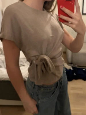Beige topp med knyt i midjan - Säljer en beige topp med rund hals och korta ärmar. Toppen har en snygg knytning i midjan som ger en cool och trendig look. Perfekt att matcha med jeans eller kjol för en avslappnad stil.