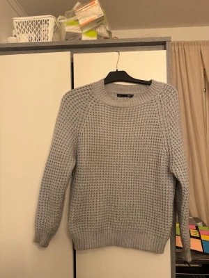 Ljusgrå stickad tröja från Lindex - Ljusgrå stickad tröja från Lindex med grovstickad struktur och rund halsringning. Tröjan har långa ärmar och ribbade muddar vid ärmslut och nederkant. Perfekt för kalla dagar och enkel att matcha med jeans eller kjol.