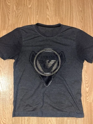 Mörkgrå t-shirt från Armani - Snygg mörkgrå t-shirt från Armani med ett stort silverfärgat logotryck framtill och mindre logga på ryggen. Klassisk rund halsringning och korta ärmar. Tillverkad i mjuk bomull för skön känsla och stilren look.
