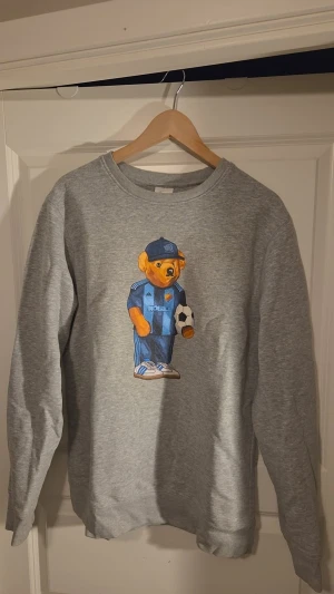 Grå sweatshirt med Djurgården-nalle - Grå sweatshirt med tryck av en söt nalle i Djurgårdens IF fotbollskläder och keps, som håller i en fotboll. Tröjan har rund halsringning och ribbade muddar vid ärmslut och nederkant. Perfekt för dig som gillar sportig och lekfull stil. Passar perfekt i vardagen men även på matchdagar!!