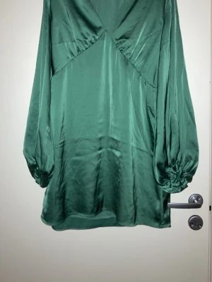 Grön omlottklänning i satin - Snygg grön kort omlottklänning i satin med långa puffärmar och v-ringning. Klänningen har en lätt glansig finish och avslappnad passform, perfekt för dig som gillar trendiga och stilrena plagg.