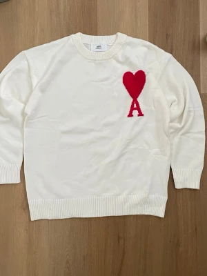 Vit stickad tröja från Ami Paris - Vit stickad tröja från Ami Paris med rund halsringning och ribbade muddar. På bröstet finns ett rött hjärta med bokstaven 'A' i samma färg. Tröjan har en avslappnad passform och är perfekt för en trendig vardagslook.