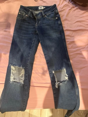 Blå slitna jeans från 157 - Ett par blå jeans från 157 med raka ben och stora slitningar på båda knäna. Klassisk femficksmodell med gylf och knapp. Jeansen har en cool, avslappnad vibe och är gjorda i denim.