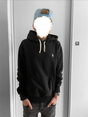 Svart hoodie från Polo Ralph Lauren - Klassisk svart hoodie från Polo Ralph Lauren med vit broderad logga på bröstet. Hoodien har huva med vita snören och en stor magficka framtill. Tillverkad i mjukt bomullsmaterial och har ribbade muddar vid ärmslut och nederkant. Priset är diskuterbart!