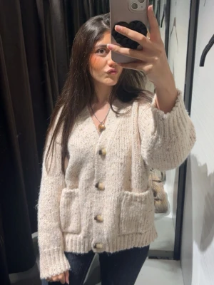 Beige stickad kofta med träknappar - Mysig beige stickad kofta med v-ringning och stora träknappar framtill. Koftan har två rejäla fickor och ribbade muddar vid ärmslut och nederkant. Perfekt för lager-på-lager och en chill look.