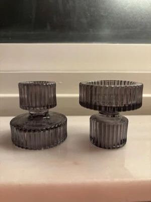 Två grå glasljusstakar - Säljer två stilrena ljusstakar i grått glas med räfflad yta och rund form. De har en modern design med olika höjd och diameter, perfekta som dekoration på bord eller fönsterbräda. Passar på båda hållen och funkar till vanliga kronljus eller doftljus och ger ett snyggt intryck i rummet.