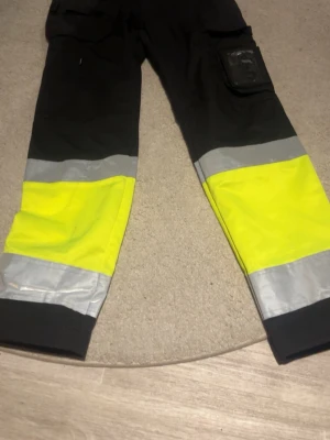 Svarta arbetsbyxor med reflexdetaljer - Arbetsbyxor i svart med stora gula och silverfärgade reflexpaneler på benen. Byxorna har flera fickor och trycket 'PRAKTISKA Gymnasiet' i orange på ena benet. Perfekta för dig som vill synas och jobba bekvämt.