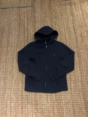 Mörkblå hoodie från Polo Ralph Lauren - Snygg mörkblå hoodie med dragkedja från Polo Ralph Lauren. Klassisk design med huva och liten röd logga broderad på bröstet. Tillverkad i mjukt bomullsmaterial och har ribbade muddar vid ärmslut och nederkant. Perfekt för en avslappnad stil. Är XL barn men sitter som xs/s