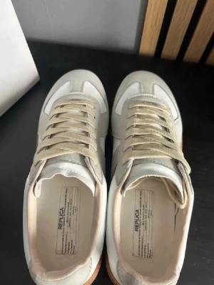 Maison Margiela gats vita - Snygga Maison Margiela Replica sneakers i beige och vitt med klassisk retrodesign. Ovandelen är i mocka och läder, med beige snören och gummisula. Perfekta för dig som gillar minimalistisk och stilren look.