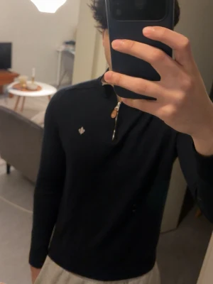 Mörkblå merinoulltröja Morris - Säljer en mörkblå stickad tröja från Morris i 100% merinoull. Tröjan har halv dragkedja med läderdetalj, liten broderad logga på bröstet och lång ärm. Perfekt för dig som gillar klassisk och stilren design.