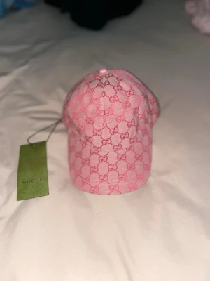 Rosa Gucci keps med monogram - Säljer en snygg rosa keps från Gucci med klassiskt GG-monogram i rött över hela kepsen. Kepsen har böjd skärm och justerbar passform. Perfekt accessoar för att lyfta din outfit med en lyxig touch.