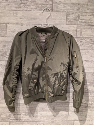 Olivgrön bomberjacka från Livelo - Snygg olivgrön bomberjacka från Livelo med dragkedja, ribbade muddar och fickor med tryckknappar. Jackan har en klassisk bombermodell med ficka på ärmen och glansigt material som ger en cool look.