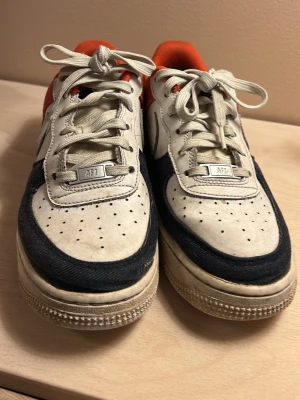 Använda Nike Air Force 1  - Nike Air Force 1 sneakers i vitt läder med jeansliknande detaljer och röd insida. Klassisk siluett med perforerad tå, vita skosnören och tydlig Swoosh-logga på sidan. Röd 'AIR'-text på sulan och metallbricka med 'AF1' på snörningen. Använda, begynnande hål vid tå. Se bild. 