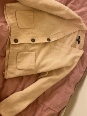 Beige kofta från Uniqlo - Mysig beige kofta från Uniqlo med knappar framtill och två stora fickor. Koftan har ribbade muddar och en avslappnad passform, perfekt att slänga på sig över en t-shirt. Tillverkad i mjukt material för extra komfort. Storlek S.