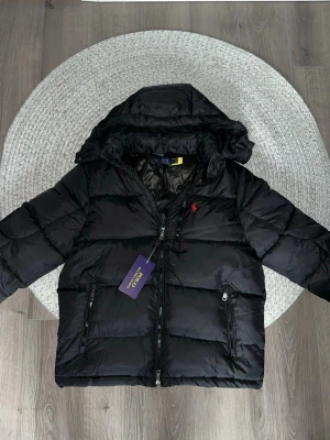 Svart pufferjacka från Ralph Lauren - Snygg svart pufferjacka från Ralph Lauren med huva och dragkedja framtill. Jackan har två fickor med dragkedja och den klassiska röda Polo-loggan på bröstet. Perfekt för kalla dagar och riktigt skön att bära.