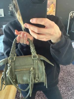 Olivgrön handväska med flätade handtag - Snygg olivgrön handväska i fejkskinn med flätade handtag och guldfärgade detaljer. Väskan har en framficka med dragkedja, dekorativa remmar och tofsar samt justerbar axelrem. Perfekt storlek för det viktigaste och ger en cool touch till din outfit.