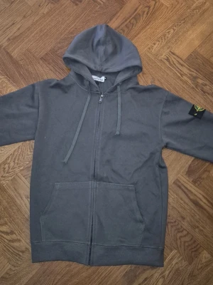 Stone Island hoodie - Säljer en mörkgrå hoodie från Stone Island med dragkedja framtill och klassisk huva med dragsko. Hoodien har en ficka på magen och den ikoniska Stone Island-loggan på vänster ärm. Perfekt för en avslappnad streetwear-look.