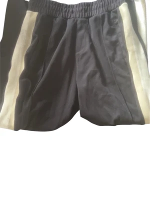 Svarta shorts med vita ränder Bershka - Säljer ett par svarta shorts från Bershka med vita sidränder. De har elastisk midja och en sportig look som passar perfekt till sommarens aktiviteter. Materialet känns mjukt och bekvämt, och designen är enkel men snygg.