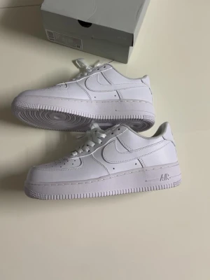 Nike Air Force 1  - Klassiska Nike Air Force 1 i helvitt läder med ikonisk swoosh på sidorna och perforerade detaljer vid tån. Tjock sula med AIR-dämpning och lågt skaft. Perfekta för en clean och stilren look.