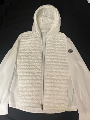  Vindjacka från Colmar  - Snygg vit vindjacka från Colmar med quiltad front, hel dragkedja och fast huva. Jackan har släta ärmar och baksida i tunt material samt Colmar-logga på ärmen. Perfekt för dig som vill ha en lätt och stilren jacka till vardags. Bara att skicka iväg ett meddelande vid frågor!