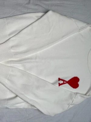 Vit stickad tröja med rött hjärta - Säljer en vit stickad tröja från AMI Paris med ett rött hjärta och bokstaven A broderat på bröstet. Tröjan har rund hals, långa ärmar och breda ribbade muddar vid ärmslut och nederkant. Perfekt för dig som gillar stilrena plagg med en twist.