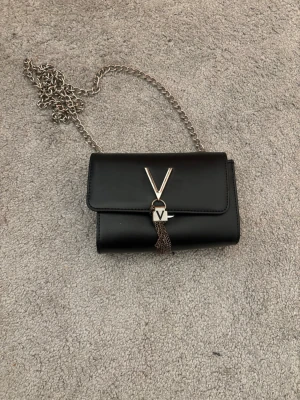 Svart handväska från Valentino - Snygg svart handväska från Valentino med silverfärgad kedja och dekorativt V-spänne framtill. Väskan har en stilren, rektangulär form och är gjord i slätt konstläder. Perfekt accessoar för att lyfta din outfit. Väskan har lite slitage, speciellt på logon, men inget man tänker på. Inköpt för 800kr