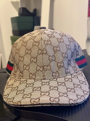 Gucci beige GG keps- komplett set - Box och allt medföljer samt kvitton!,         Säljer en beige keps från Gucci med klassiskt GG-monogram över hela kepsen. Kepsen har gröna och röda ränder på sidorna och justerbart spänne bak i läder. Materialet är canvas och detaljerna är i brunt. Perfekt för dig som gillar exklusiv streetwear.