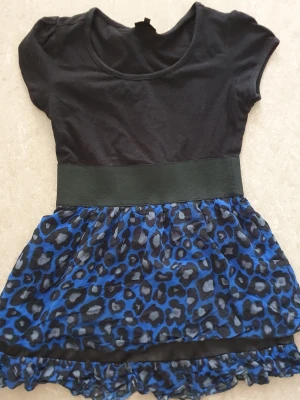 Klänning - Klänning från KappAhl med svart överdel i mjuk bomull och korta ärmar. Kjoldelen har blått och svart leopardmönster i lätt, luftig polyester med volangkant nertill och bred midjeresår. Perfekt för dig som gillar djurmönster och färg!