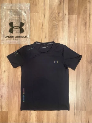 Svart tränings-tshirt från Under Armour - Svart t-shirt från Under Armour med korta ärmar och rund hals. Tillverkad i ett lätt och stretchigt syntetmaterial, perfekt för träning. Diskret logga på bröstet och tryck på ärmen samt sidan. Snygg och sportig design.