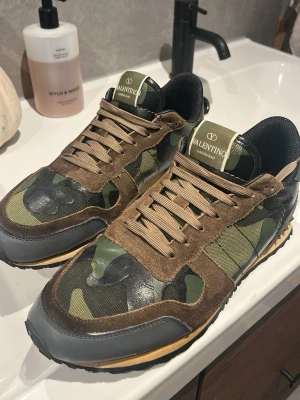 Valentino camo sneakers med mockadetaljer - Snygga sneakers från Valentino Garavani med grön camo-mönstrad textil, bruna mockadetaljer och svarta läderpartier. Skorna har beige snören, svart innersula och cool gummisula med nitar på hälen. Perfekta för dig som vill sticka ut med stil. Box och skopåse ingår, tveka inte på att höra av dig vid funderingar!