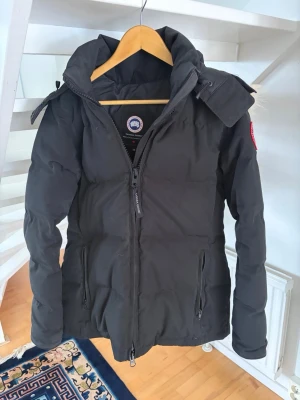 Svart dunjacka från Canada Goose - Säljer en svart dunjacka från Canada Goose med klassisk logotyp på ärmen och avtagbar huva med pälsdetalj. Jackan har två dragkedjeförsedda fickor och hög krage som skyddar mot kyla. Perfekt för kalla vinterdagar.