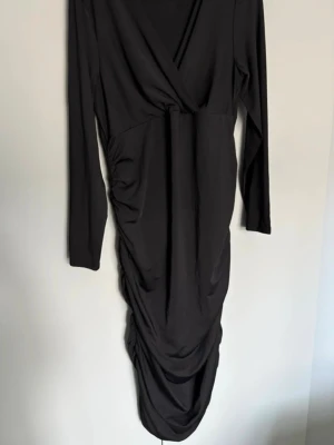 Svart omlottklänning med drapering 42  - Svart långärmad klänning med omlott-ringning och snygg drapering längs sidan och framtill. Klänningen har en elegant, figurnära passform och är tillverkad i ett mjukt, stretchigt material som ger en bekväm känsla. Ny utan pris lapp