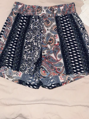 Mönstrade shorts från Jacqueline de Yong - Säljer ett par snygga shorts från Jacqueline de Yong med färgglatt paisley- och blommönster i blått, vitt och rött. De har elastisk midja och är tillverkade i ett mjukt, tunt material som passar perfekt till varma dagar.