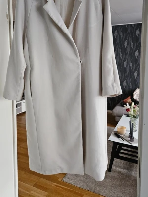 Ljusbeige lång kappa med enkel knäppning - Stilren ljusbeige kappa i klassisk rak modell med enkel knäppning framtill och långa ärmar. Kappan har en snygg krage och är tillverkad i ett mjukt tyg som ger en elegant look. Perfekt för dig som gillar minimalistisk och tidlös stil. Tunt foder, lite kraftigare tyg utanpå. Vinter/ vår kappa oversize