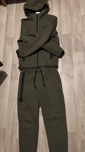 Grön nike tech tracksuit - Säljer en olivgrön hoodie från Nike med hel dragkedja framtill och två fickor med svarta dragkedjor. Hoodien har en klassisk huva och diskret Nike-logga på bröstet. Materialet är mjukt och stretchigt, perfekt för en avslappnad streetwear-look.