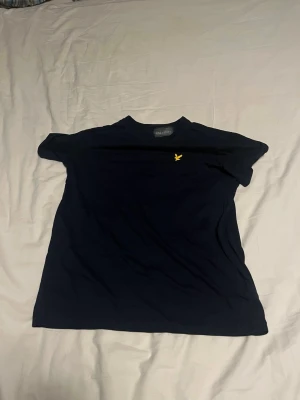 Mörkblå t-shirt från Lyle & Scott - Snygg mörkblå t-shirt från Lyle & Scott med klassisk gul logga på bröstet. T-shirten har rund hals och korta ärmar, perfekt för en clean och stilren look. Tillverkad i mjuk bomull som känns skön mot huden.