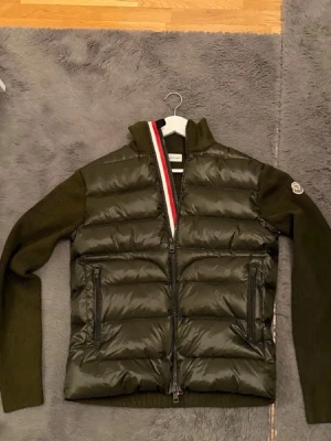 Olivgrön dunväst från Moncler ÄKTA!!  -   Säljer billigt för den inte passar mig JA ALLA QR KODER OCH SCANS FUNGERAR Snygg olivgrön dunväst från Moncler med stickade ärmar och quiltad front. Västen har dragkedja, två fickor fram och Moncler-logga på ärmen. Detaljer i rött, vitt och blått längs dragkedjan ger en sportig touch. Perfekt för lager-på-lager och streetstyle.