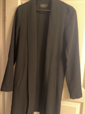 Svart lång kappa från ONLY - Stilren svart kappa från ONLY med rak passform och öppen front. Kappan har långa ärmar, två diskreta fickor framtill och en klassisk krage. Materialet ser ut att vara en mjuk och slät textil som passar perfekt till höst och vår. Finns knytband till.