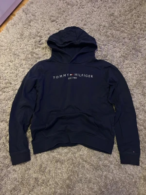Marinblå hoodie från Tommy Hilfiger - Snygg marinblå hoodie från Tommy Hilfiger med klassisk logga och texten 'EST. 1985' på bröstet. Hoodien har huva och en stor magficka framtill. Tillverkad i mjukt bomullsmaterial och har ribbade muddar vid ärmslut och nederkant. Size: 164cm