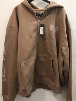 Brun set med hoodie och  mjukisbyxor från BoohooMAN - Säljer ett par beige mjukisbyxor och hoodie med dragkedja från BoohooMAN med resår och dragsko i midjan. Byxorna är i mjukt bomullsmaterial och har en avslappnad passform, perfekta för chill dagar. Matcha med hoodie för en komplett look.