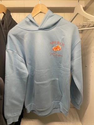 Ljusblå Aperol hoodie med tryck - Ljusblå hoodie från Aperol med stort tryck av två glas Aperol Spritz och texten 'Spritz Happens' på bröstet och ryggen. Hoodien har känguruficka, huva och ribbade muddar. Perfekt för dig som gillar snygga och chill plagg med coolt motiv.