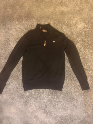 Morris halvzip svart - Snygg svart stickad Morris tröja med halvzip och liten broderad logga på bröstet. Tröjan har lång ärm, ribbade muddar och hög krage med dragkedja. Perfekt för lager på lager och enkel att matcha med olika outfits.