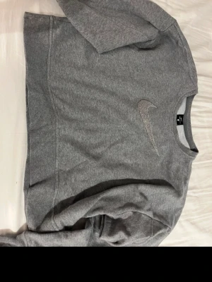Grå Nike Dri-Fit sweatshirt - Säljer en grå sweatshirt från Nike med Dri-Fit material. Tröjan har rund hals, långa ärmar och en stor Nike Swoosh-logga broderad på bröstet. Perfekt för en sportig och avslappnad stil.