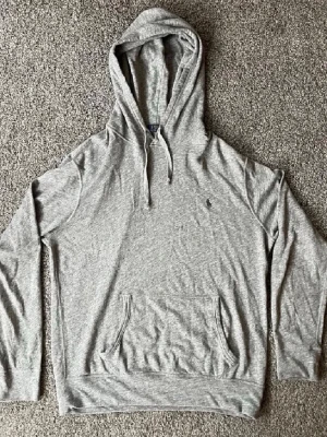 Grå hoodie från Polo Ralph Lauren - Säljer en stilren grå hoodie från Polo Ralph Lauren med klassisk huva och dragsko. Hoodien har en känguruficka framtill och den ikoniska lilla loggan broderad på bröstet. Tillverkad i mjuk bomullsblandning som är skön att bära hela dagen. Väldigt bra skick förutom ett litet hål, be gärna om bild