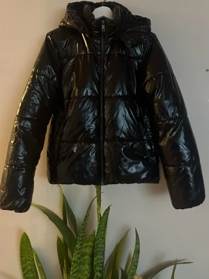 Svart glansig pufferjacka med huva - Jättefin svart jacka från Nelly. Toppenskick förutom en defekt på högra armen syns knappt men man kan säkert fixa med lappning eller något. 